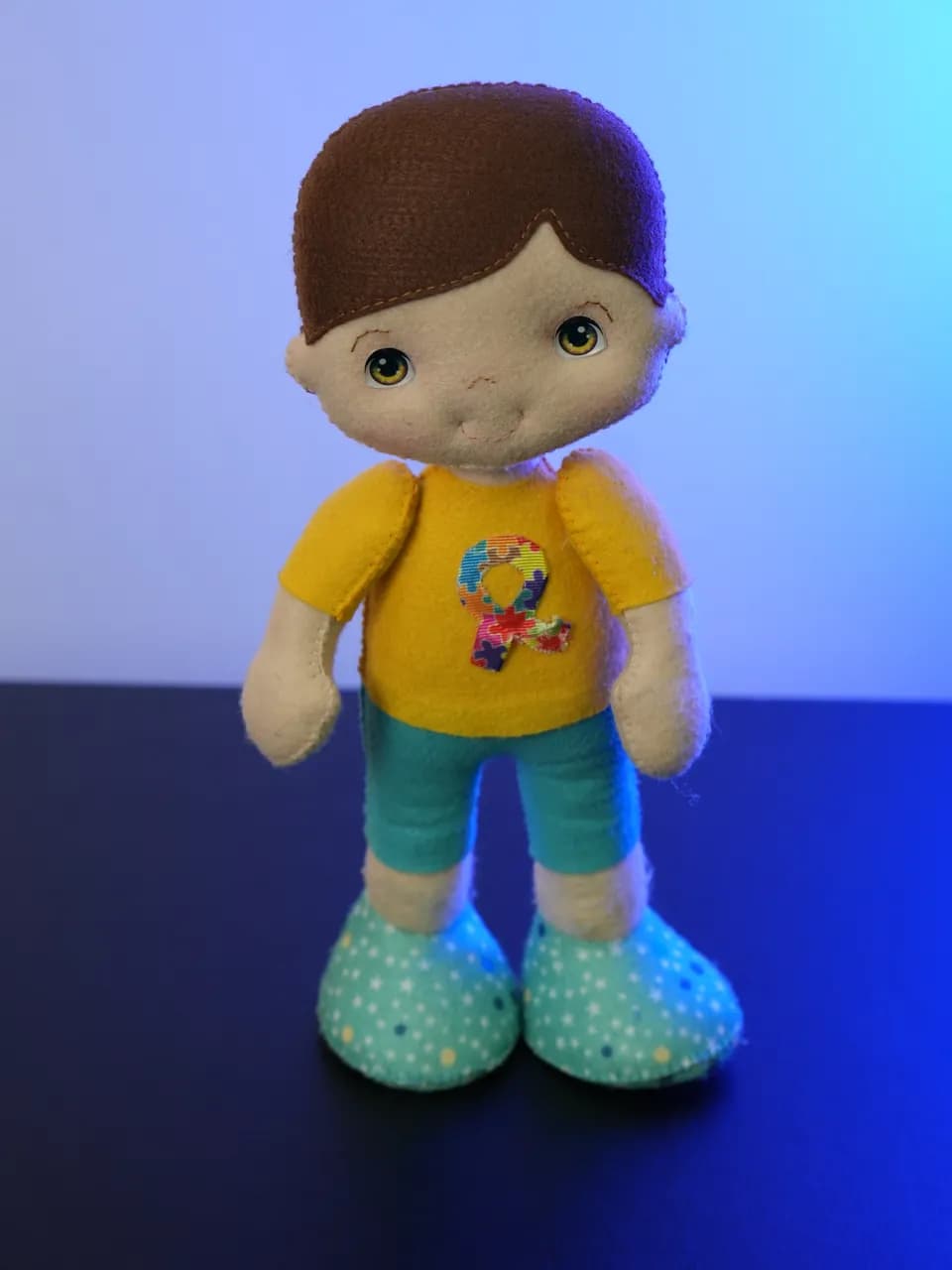 O Dom: boneco de feltro de cabelo castanho, camiseta amarela com um laço colorido no peito e bermuda azul