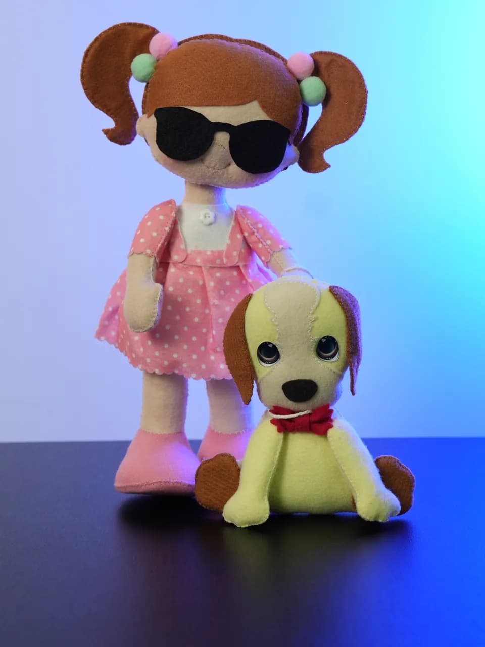 A Nina: boneca de feltro de maria-chiquinha e óculos escuros, vestido rosa de bolinhas, ao lado do cão-guia de feltro amarelo