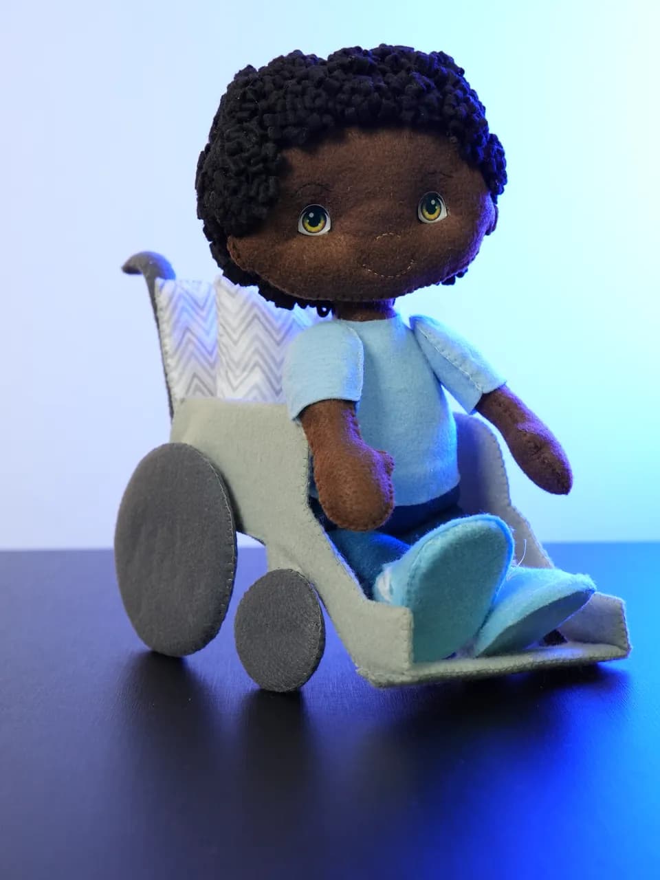 O Theo: boneco de feltro de cabelo crespo preto e camisa azul, sentado na cadeira de rodas de feltro