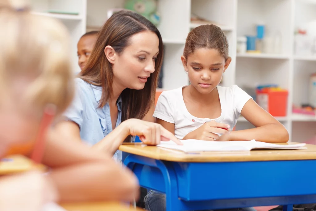 Uma professora sentada ao lado de uma aluna em sala de aula aponta para um caderno aberto sobre a mesa, orientando a atividade. Outras crianças aparecem ao fundo, em um ambiente escolar organizado e iluminado.
